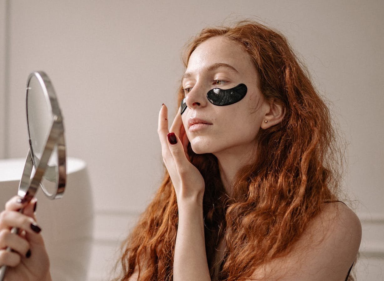 Tjej med eye patches under ögonen som speglar sig i en liten spegel.